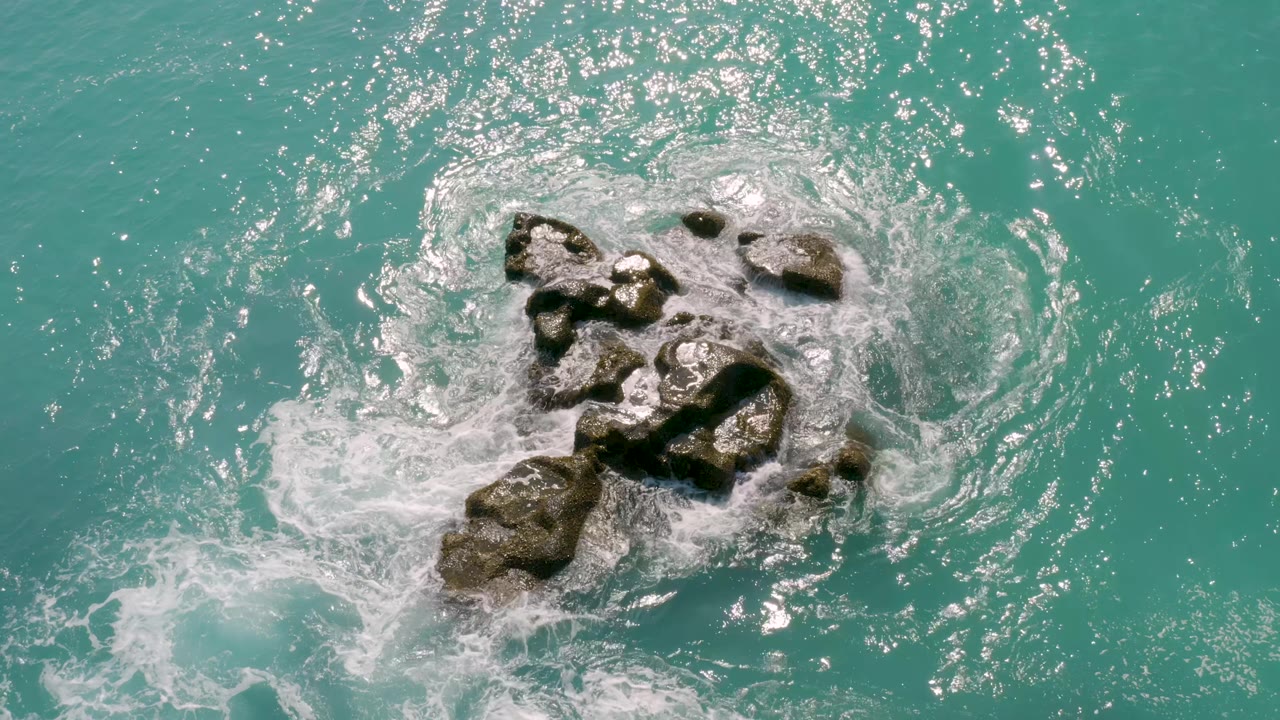 Aerial frame from Dreams Beach with a thicker band of white surf spreading over the left side of the rock cluster in clear blue water, with no visible people.