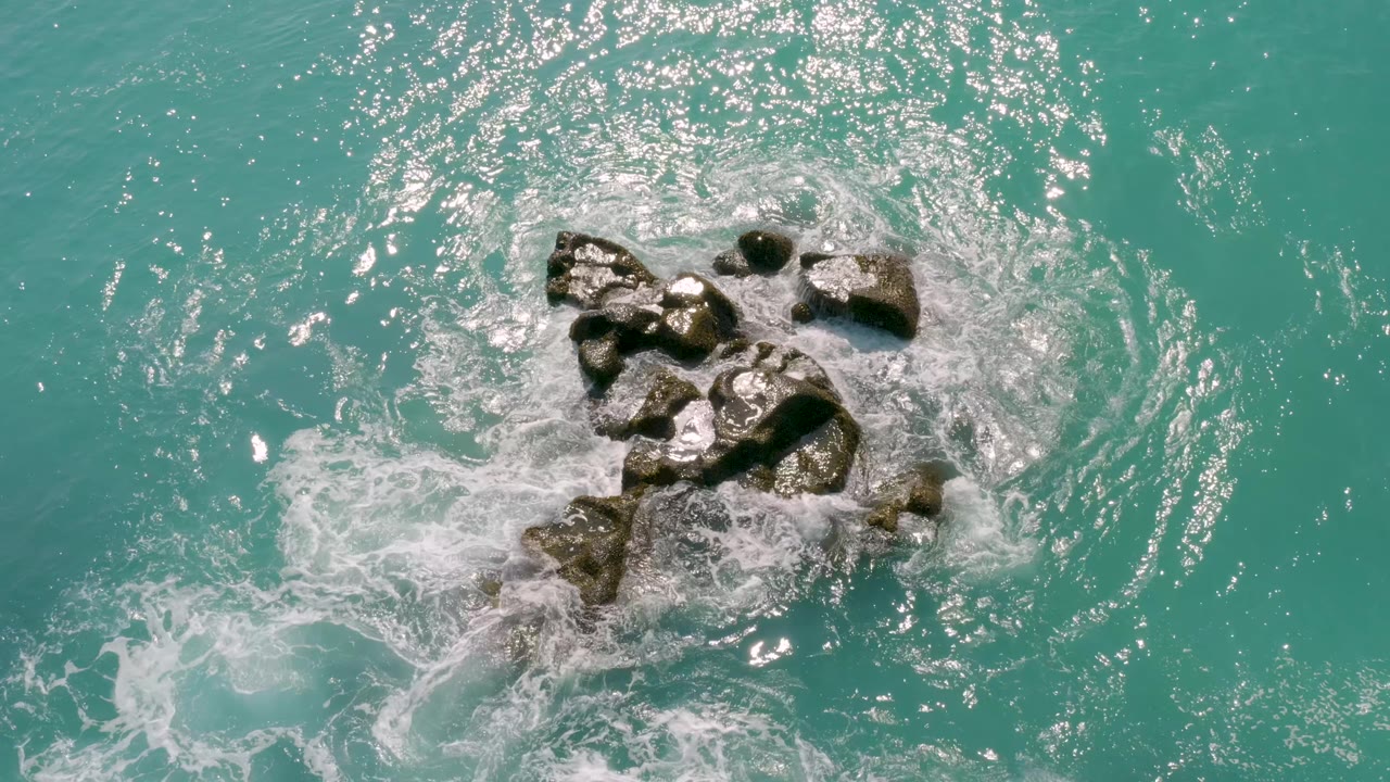 Overhead Dreams Beach view where white foam curls around the upper rocks and bright reflections scatter across the turquoise water, with no visible people.