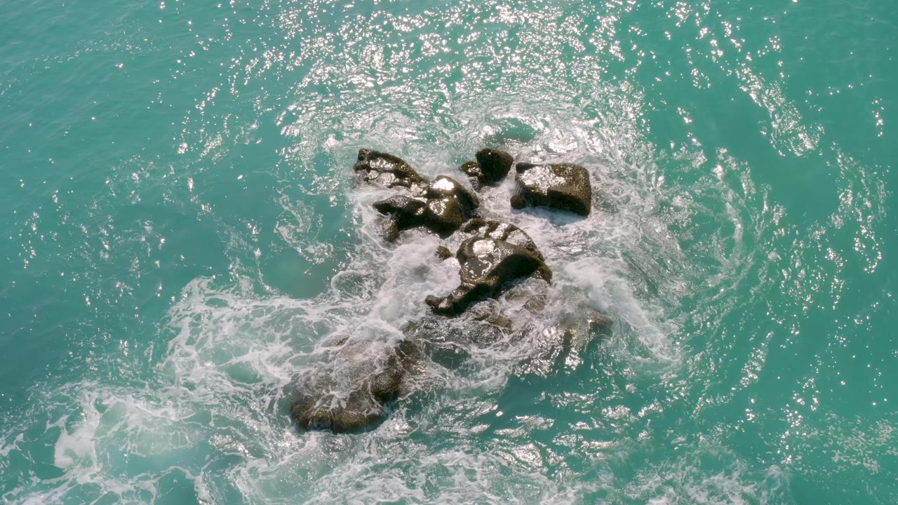 Aerial view in Baja California Sur with the rock cluster slightly lower in frame as pale foam thins across the surrounding clear sea, with no visible people.