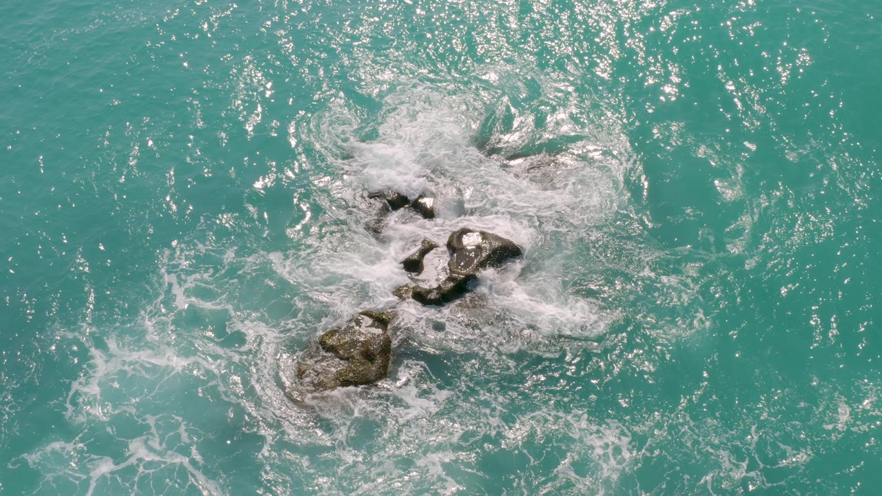 Overhead view at Dreams Beach in Baja California Sur, Mexico, with white surf breaking around a cluster of dark rocks in turquoise water and no visible people.