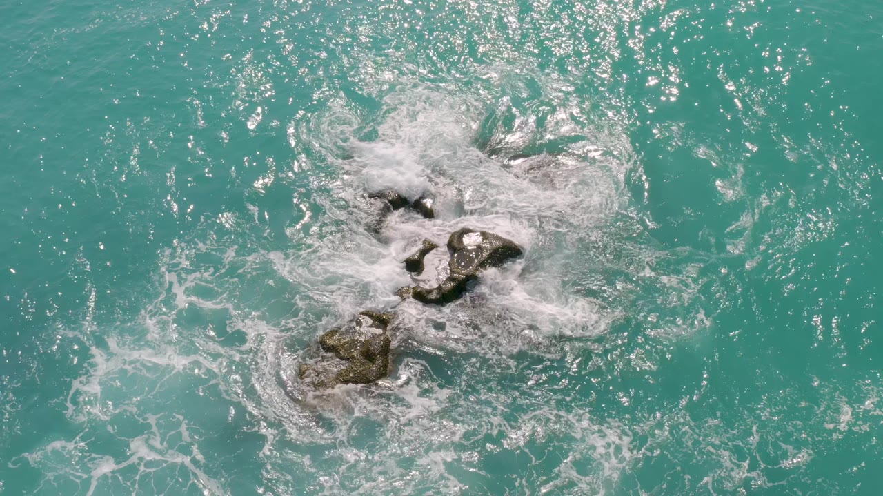 Aerial frame from Dreams Beach in Baja California Sur, Mexico, where foamy water converges from multiple sides around exposed rocks, with no visible people.