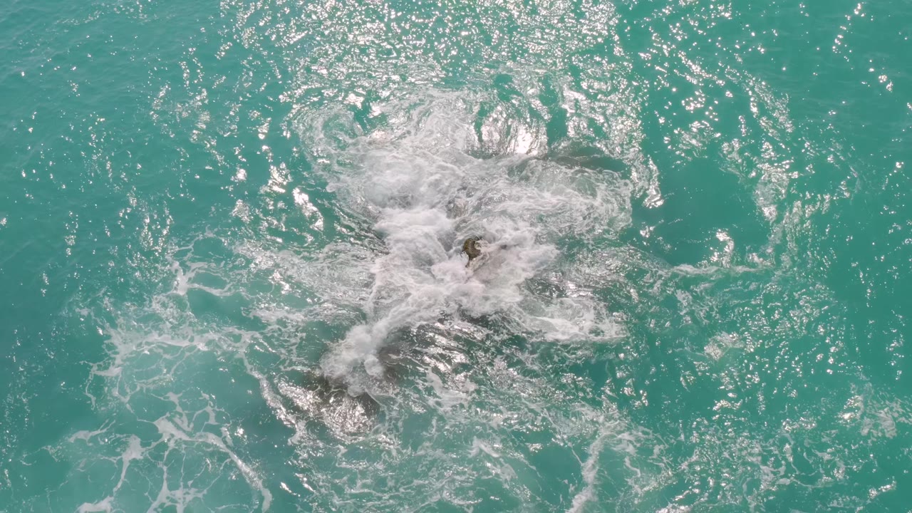 Overhead ocean view at Dreams Beach in Baja California Sur, Mexico, with white surf spreading over most of the central rock cluster in turquoise water and no visible people.