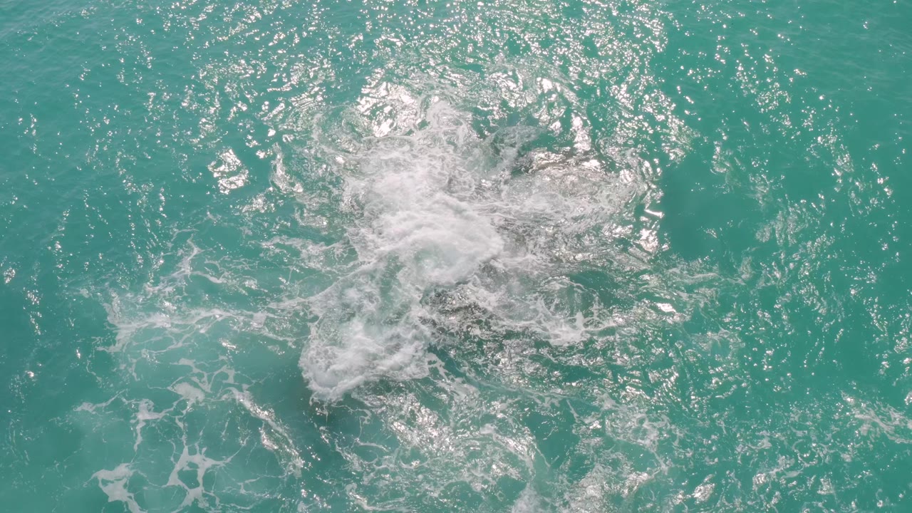 A foamy white plume drifts across turquoise water at Dreams Beach in Baja California Sur, Mexico, leaving only faint dark rock shapes visible below or within the surf, with no visible people.