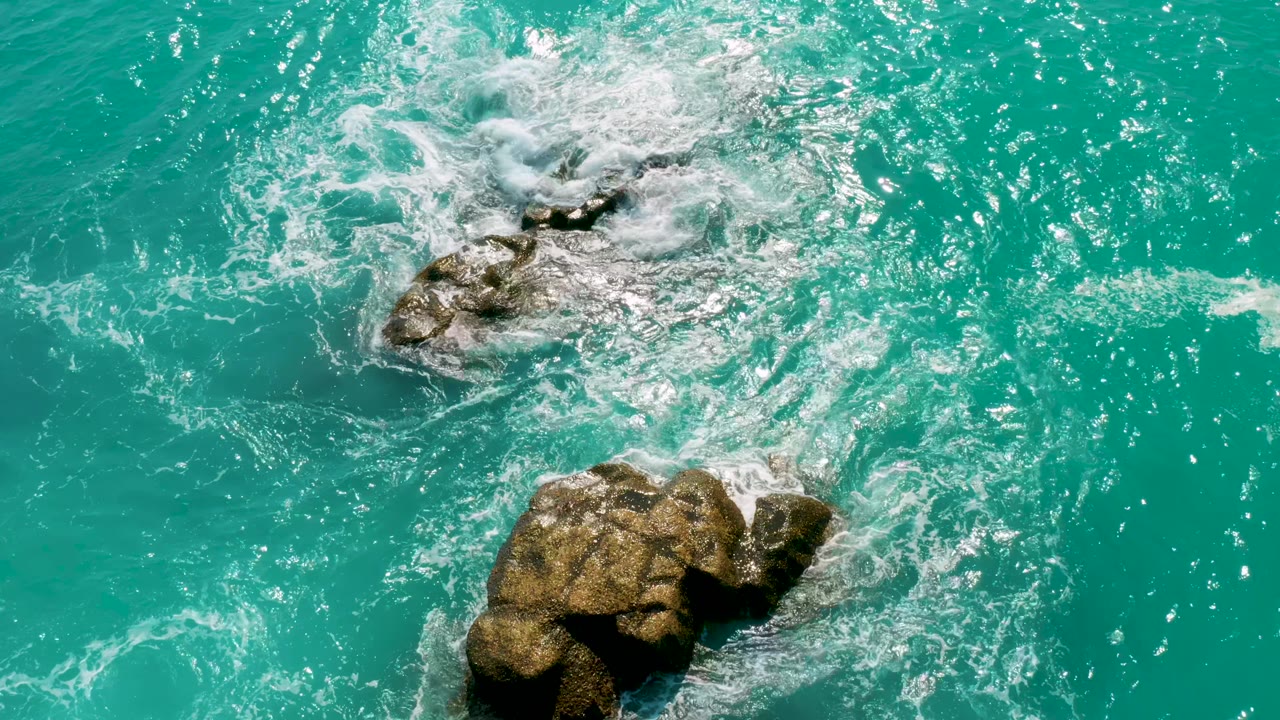 At 0:01, white foam spreads farther across the upper rock while a larger brown rock sits lower in the turquoise water at Dreams Beach, with no visible people.