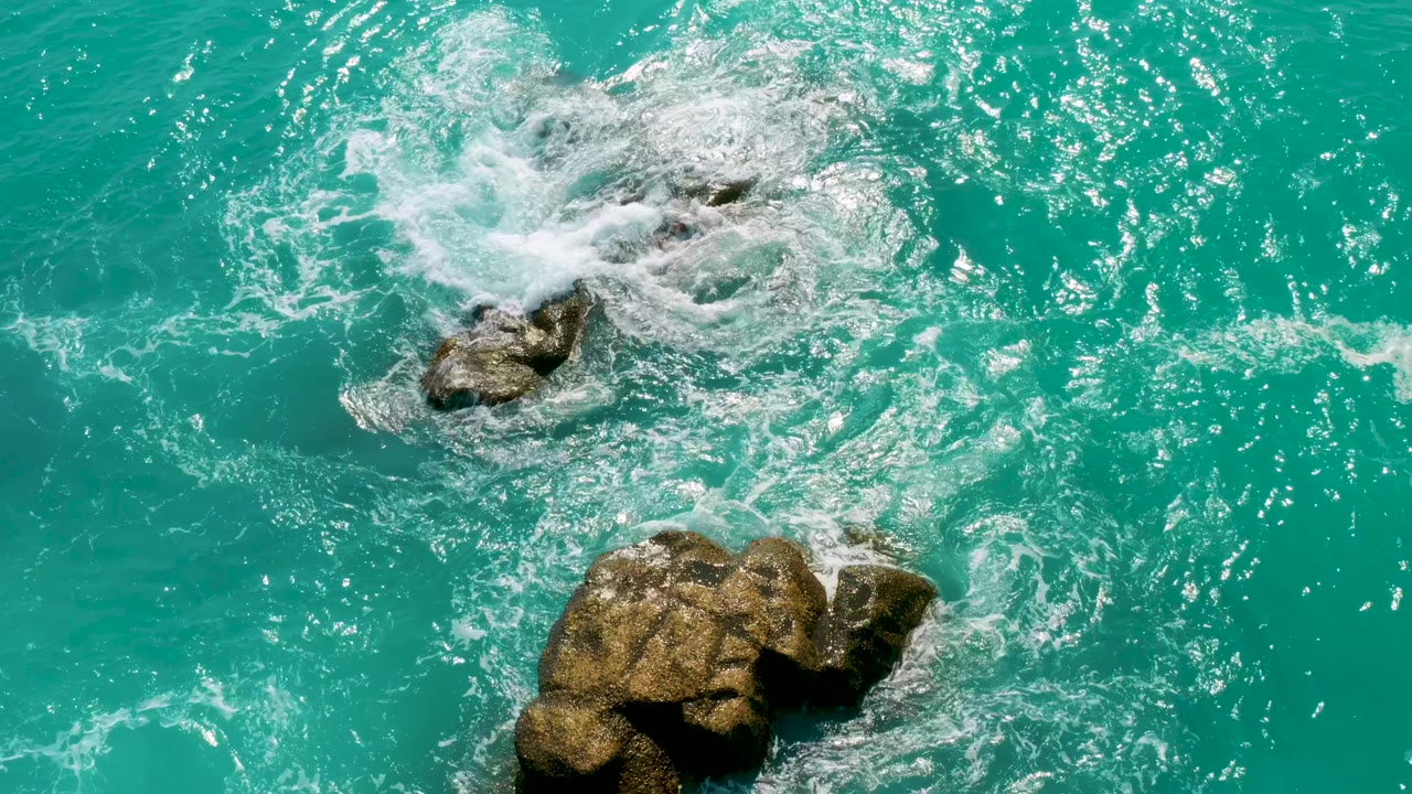 White surf curls across the top rock group while the lower rock remains partly surrounded by clear blue-green water at Dreams Beach, Baja California Sur, Mexico, with no visible people.