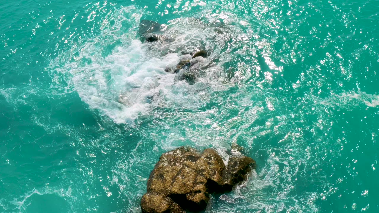 Mid-clip, a larger patch of white wash covers the upper rocks while the lower rock sits near the bottom edge in bright turquoise water, with no visible people.