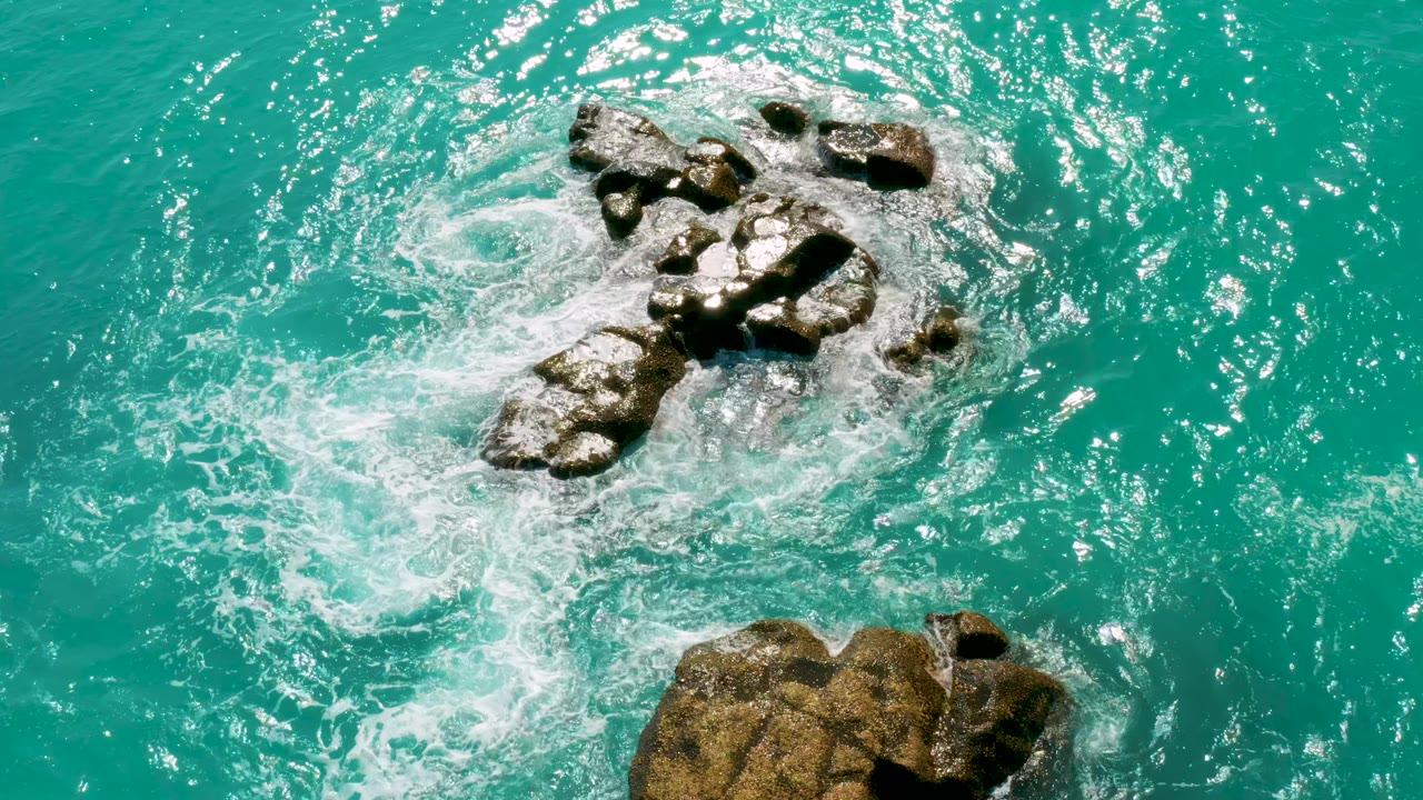 Aerial-style frame at Dreams Beach in Baja California Sur shows white foam spreading from a rocky outcrop into bright turquoise water, with no visible people.