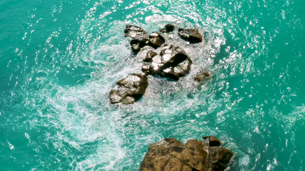Overhead frame at Dreams Beach in Baja California Sur shows exposed rocks near the top of frame and a separate rock edge below, surrounded by sunlit turquoise water, with no visible people.