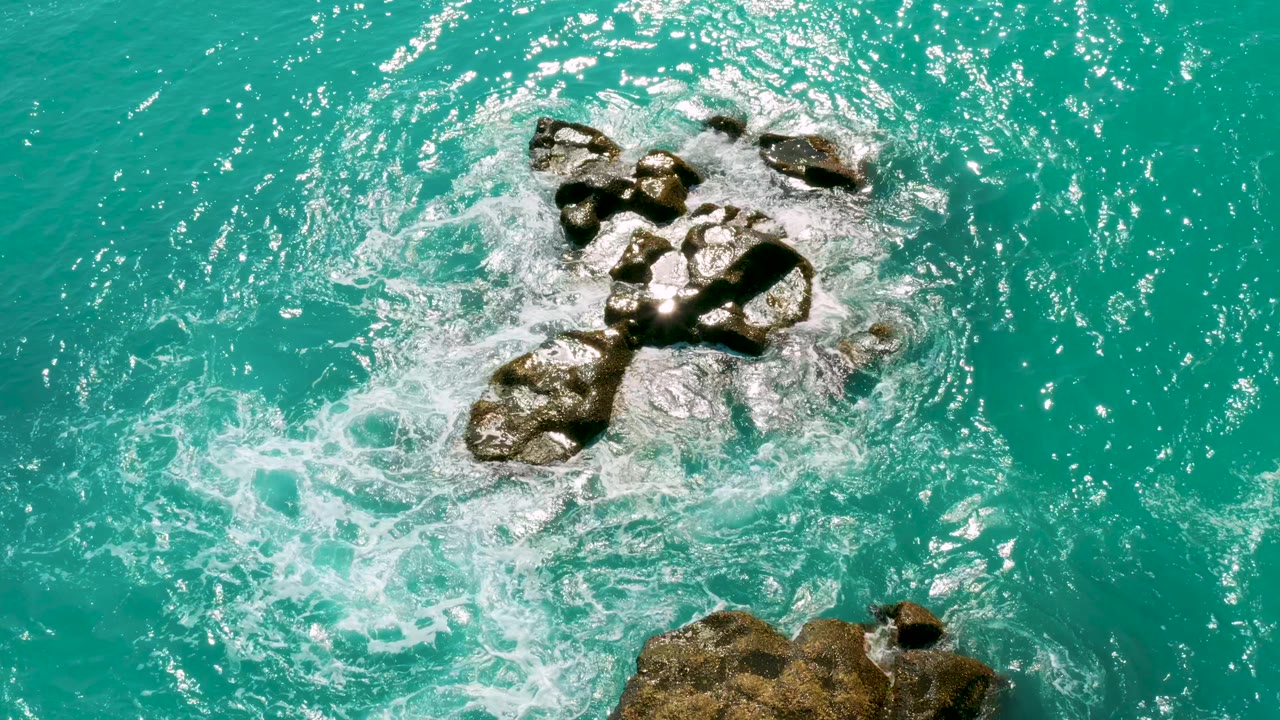 A high overhead view at Dreams Beach in Baja California Sur shows foamy water crossing dark rocks in clear blue sea, with no visible people.