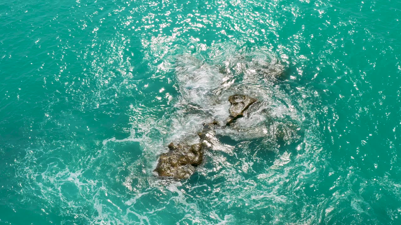 Overhead view at Dreams Beach, Baja California Sur, Mexico, with turquoise water and white foam surrounding a brown rock cluster, no visible people.