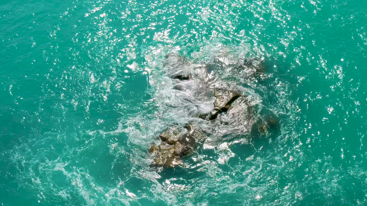 The rock cluster sits slightly right of center as foam curls around its upper edge in clear green-blue water at Dreams Beach, no visible people.