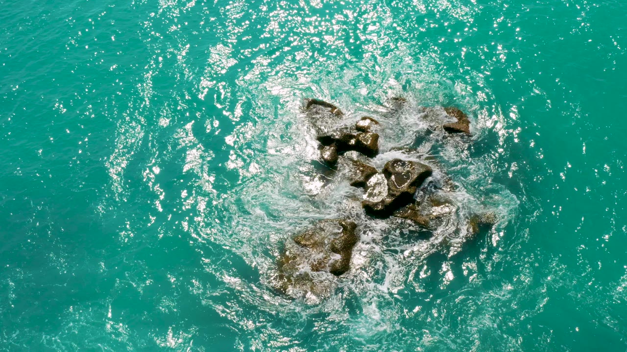 Overhead view of dark rocks surrounded by turquoise water and white foam at Dreams Beach in Baja California Sur, Mexico, with no visible people.