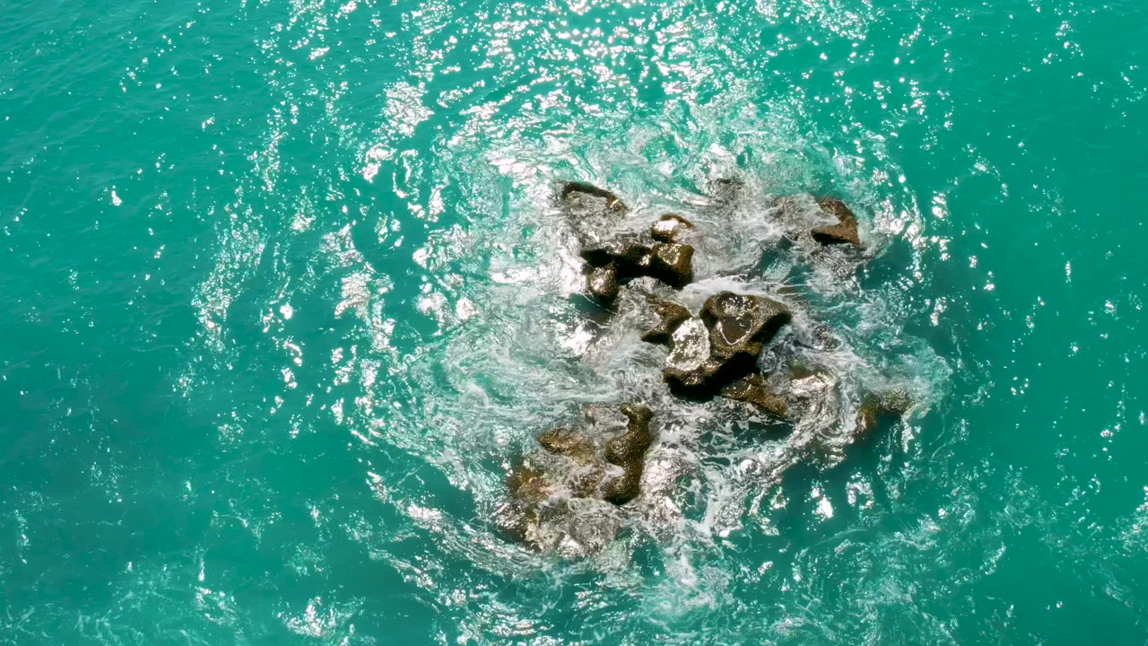 Aerial frame of turquoise sea water circling a central rock cluster at Dreams Beach in Baja California Sur, Mexico, with bright sun reflections and no visible people.