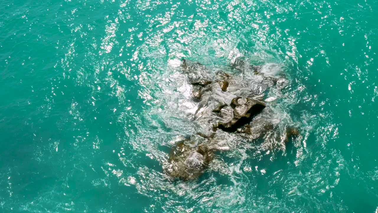 Top-down view of white surf spreading over exposed rocks in turquoise water at Dreams Beach in Baja California Sur, Mexico, with no visible people or structures.