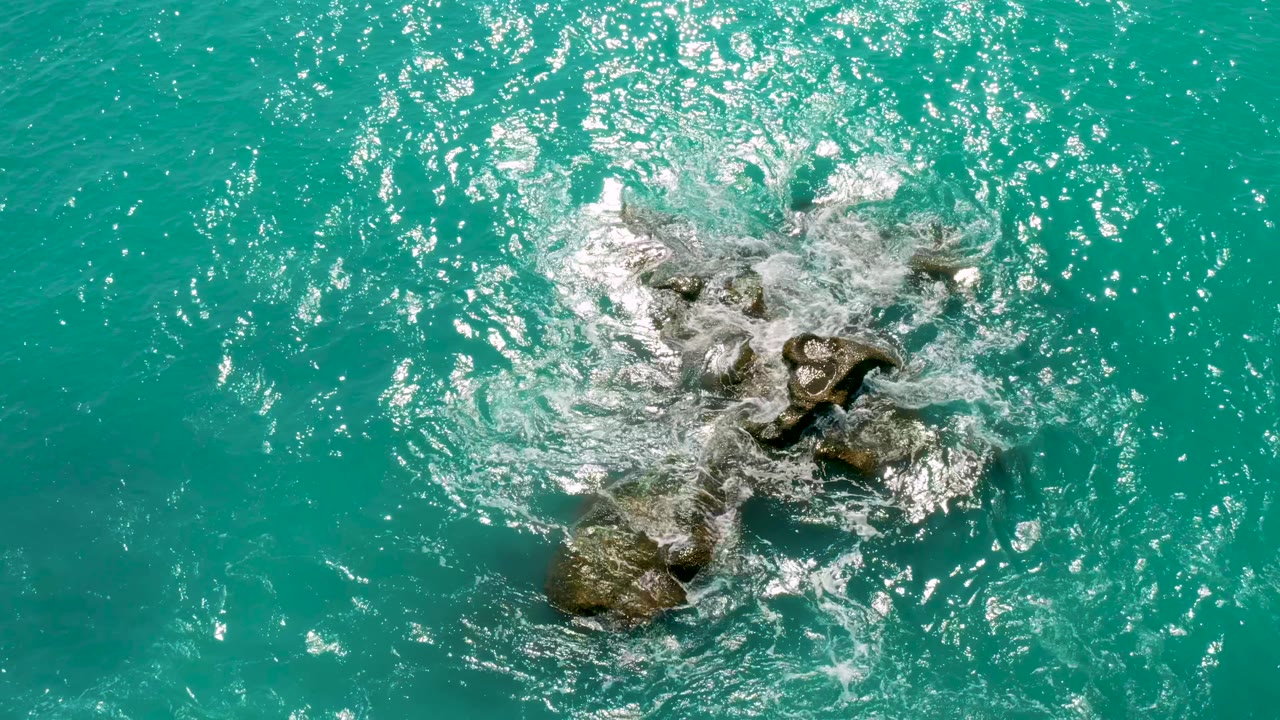 Overhead coastal frame where foam fans across dark rocks in turquoise sea at Dreams Beach in Baja California Sur, Mexico, with no visible people.