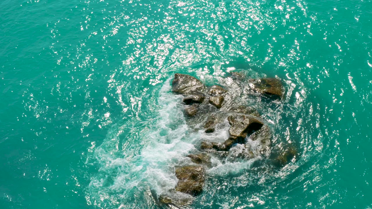 Top-down view at Dreams Beach in Baja California Sur as turquoise water foams around a cluster of wet rocks near the center of frame, with no visible people.