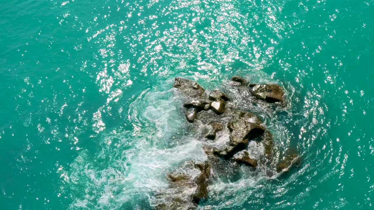 Overhead coastal frame from Dreams Beach in Baja California Sur showing white foam curling around dark rocks, open turquoise water, and strong sun sparkle across the upper frame, with no visible people.