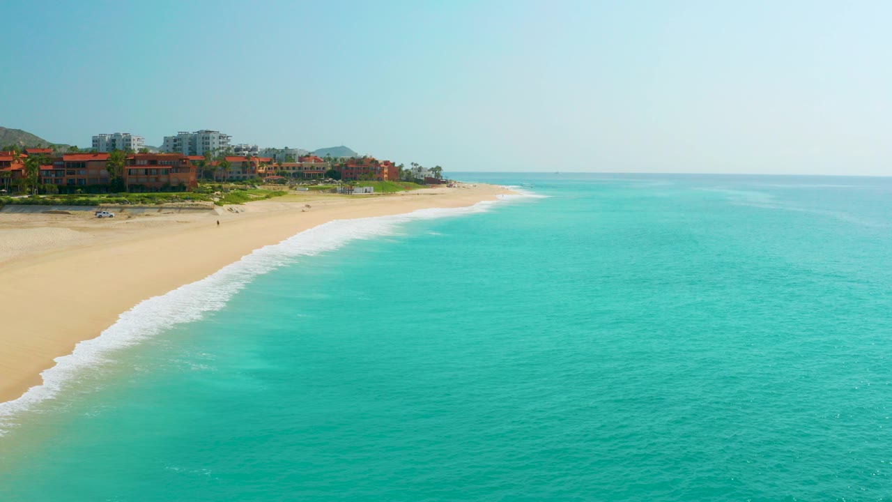 Aerial view at Dreams Beach in Baja California Sur, Mexico, with turquoise sea in the foreground, white surf along the sand, coastal buildings to the left, and no visible people.