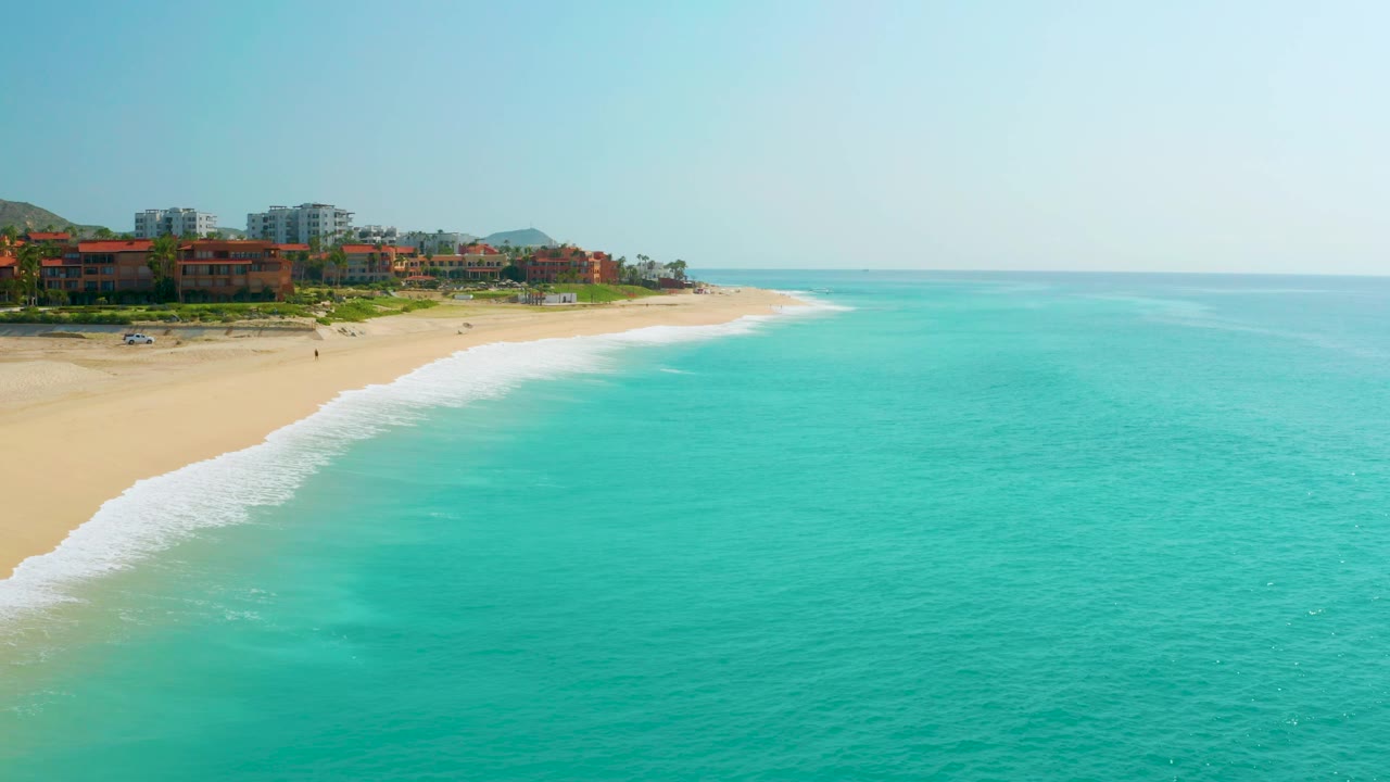 Aerial view of Dreams Beach in Baja California Sur, Mexico, with open turquoise water dominating the frame, a sandy beach on the left, distant buildings, and no visible people.