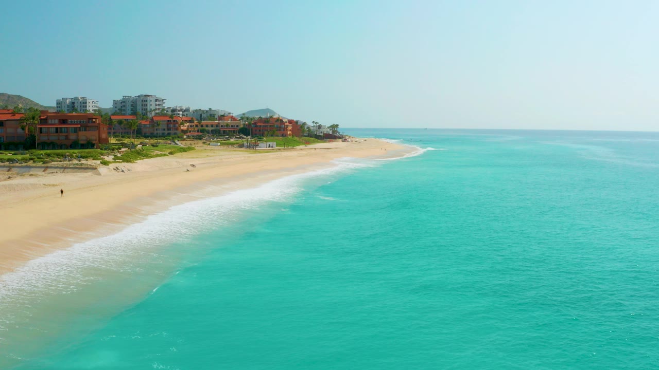 Opening aerial frame over Dreams Beach in Baja California Sur, Mexico, with turquoise water on the right, pale sand on the left, coastal buildings in the distance, and no visible people.