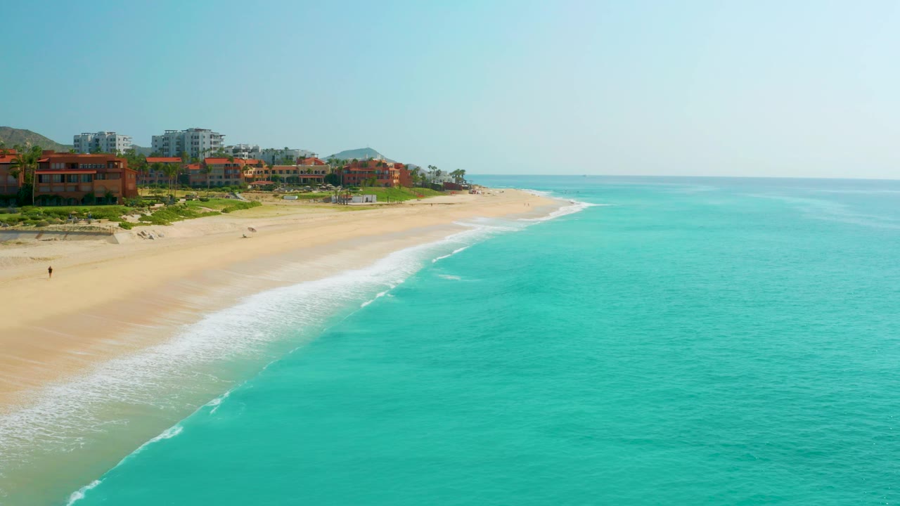 Drone-style frame over Dreams Beach showing blue-green ocean, a long pale beach, low red-orange buildings, taller background buildings, and no visible people.