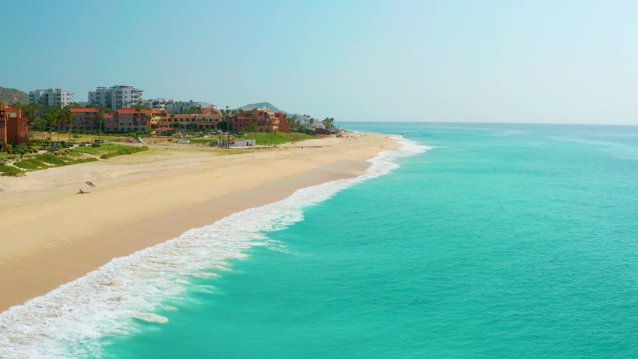 Opening aerial frame of Dreams Beach in Baja California Sur, Mexico, with resort buildings at left, a wide sandy beach, turquoise surf, and no visible people.