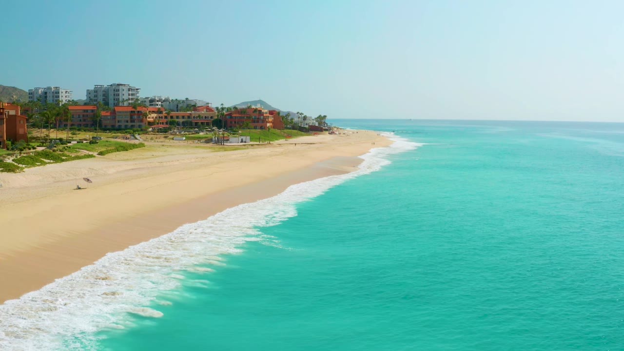 Early frame of Dreams Beach in Baja California Sur, Mexico, showing the shoreline angling from resort buildings toward bright turquoise water, with no visible people.