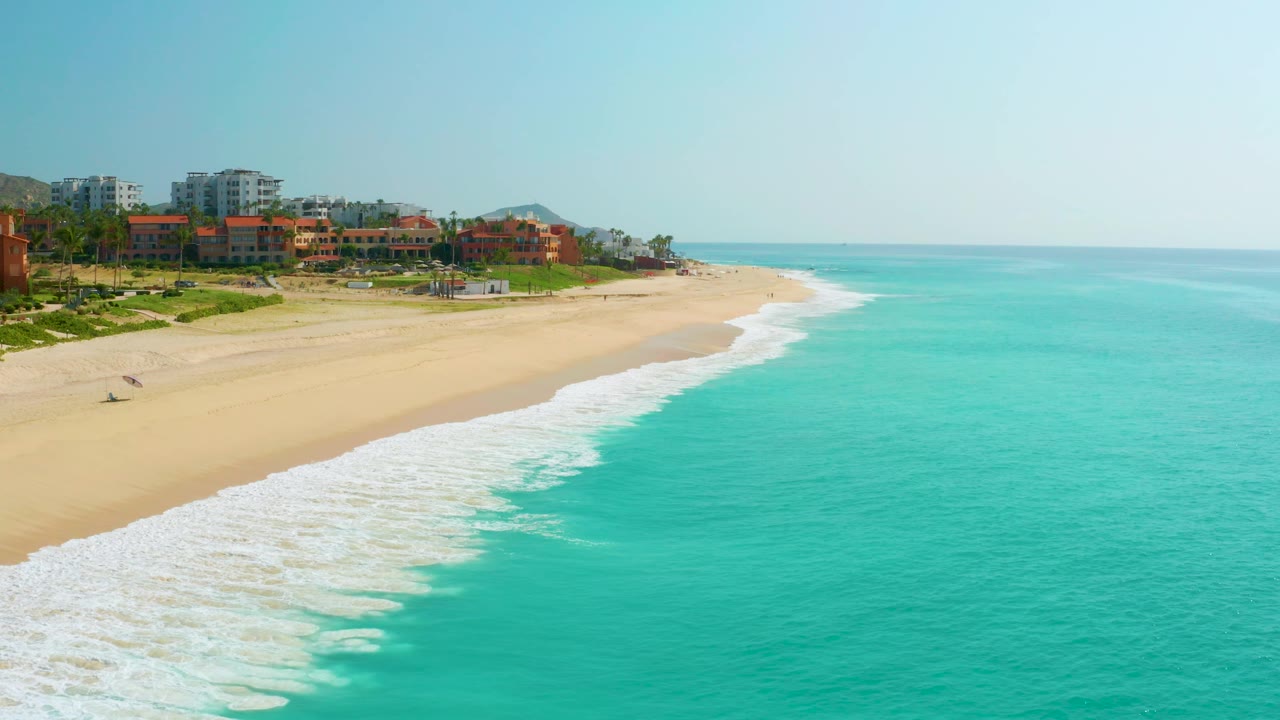 Aerial view over Dreams Beach in Baja California Sur, Mexico, with more open ocean on the right, resort structures on the left, a foamy shore break, and no visible people.