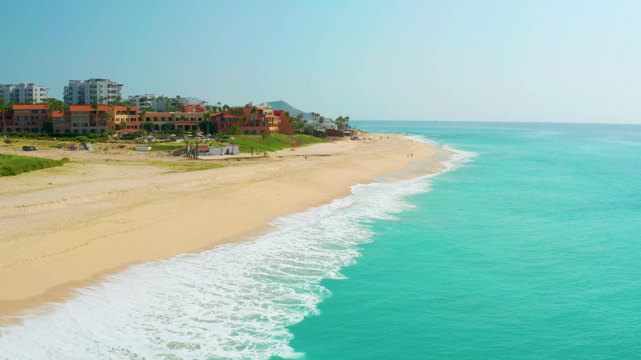 At 0:02, the coastline curve is more centered, with white foam spreading along the sand, clear blue water filling the right half, Baja California Sur beachfront buildings on the left, and no visible people.