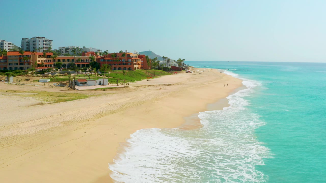 Opening aerial frame over Dreams Beach in Baja California Sur, with turquoise water rolling onto broad sand and orange-roofed buildings along the left edge.