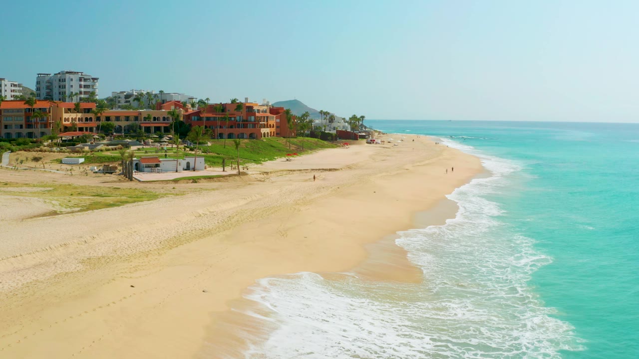 Dreams Beach aerial frame showing light blue water, a long open strip of sand, and clustered orange-roofed buildings set back from the shore.