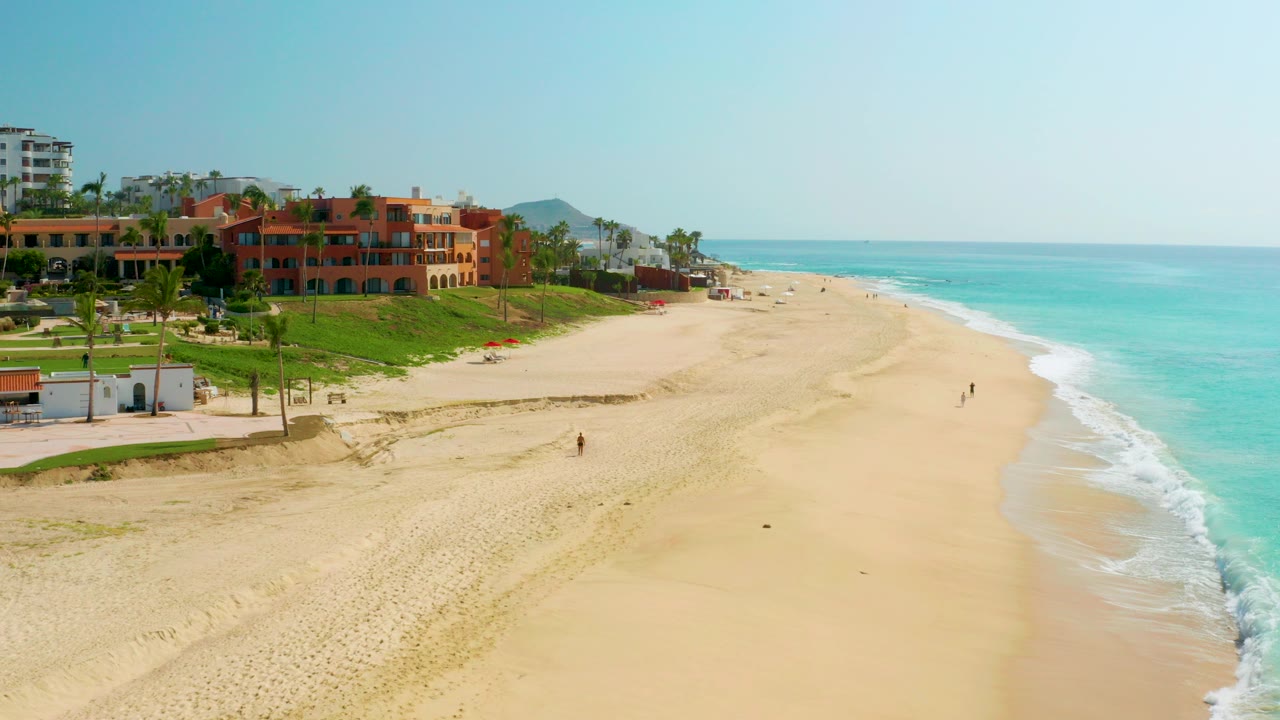 Wide aerial frame at Dreams Beach, Baja California Sur, showing pale sand, turquoise water, red-orange beachfront buildings, palms, and small distant people on the beach.