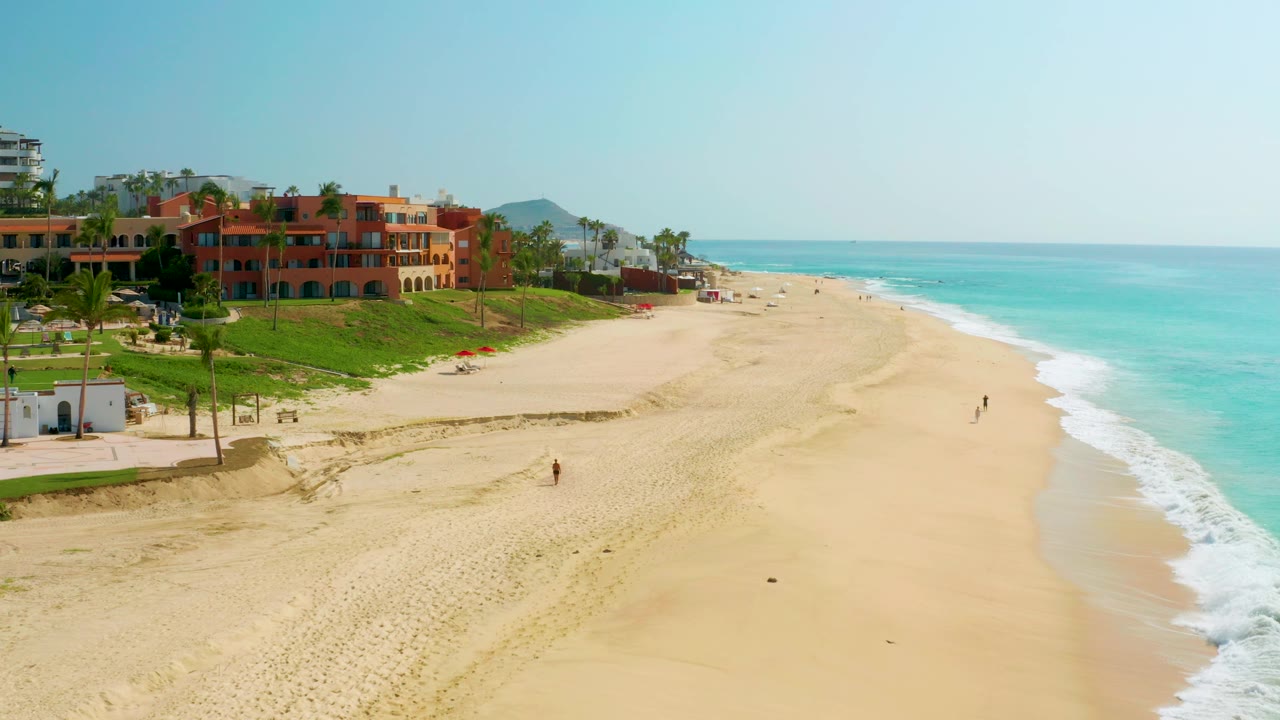 Aerial frame at Dreams Beach with red-orange buildings and green landscaping on the left, broad sand through the center, blue ocean on the right, and distant beachgoers visible.
