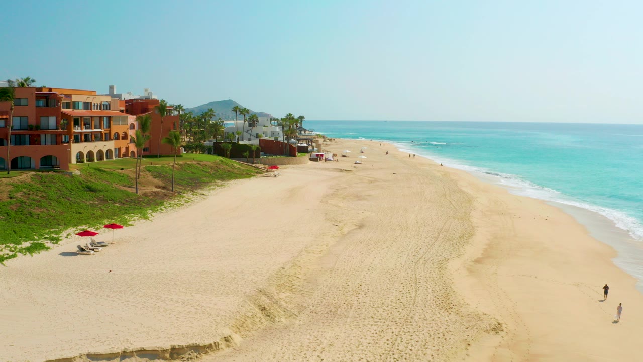 Wide aerial view of Dreams Beach in Baja California Sur with beachfront buildings at left, broad golden sand, turquoise water at right, and small distant beachgoers near the shore.