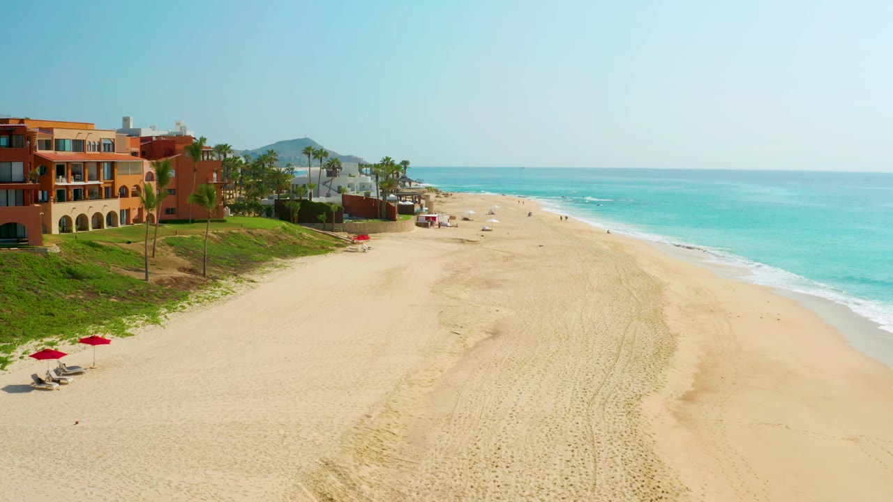 Aerial view of Dreams Beach with orange beachfront architecture at left, palms behind a green dune edge, raked sand across the center, and pale turquoise water under a clear sky.