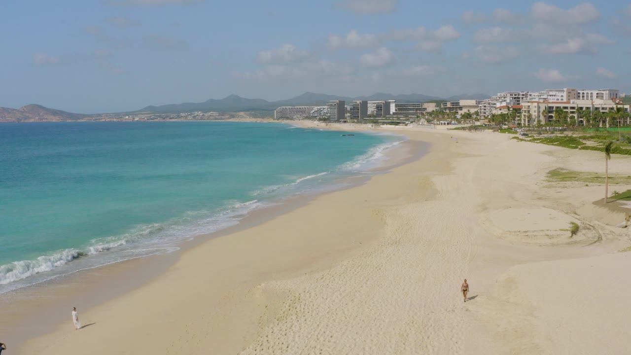At Dreams Beach in Baja California Sur, turquoise water curves into a wide pale beach with distant oceanfront buildings, hazy hills, and tiny beachgoers near the lower edge.