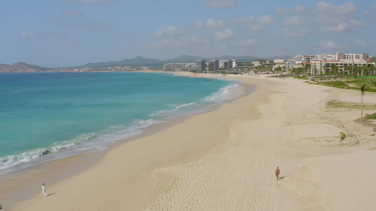Aerial beach view with blue water on the left and a broad sandy shoreline curving toward buildings.