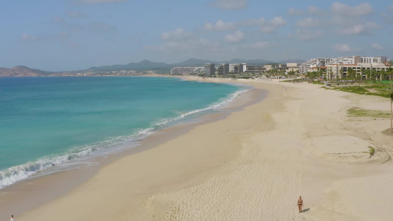 At Dreams Beach, a band of foamy surf reaches farther up the pale sand while turquoise water, resort-style oceanfront buildings, palms, and distant hills remain visible in bright daylight.
