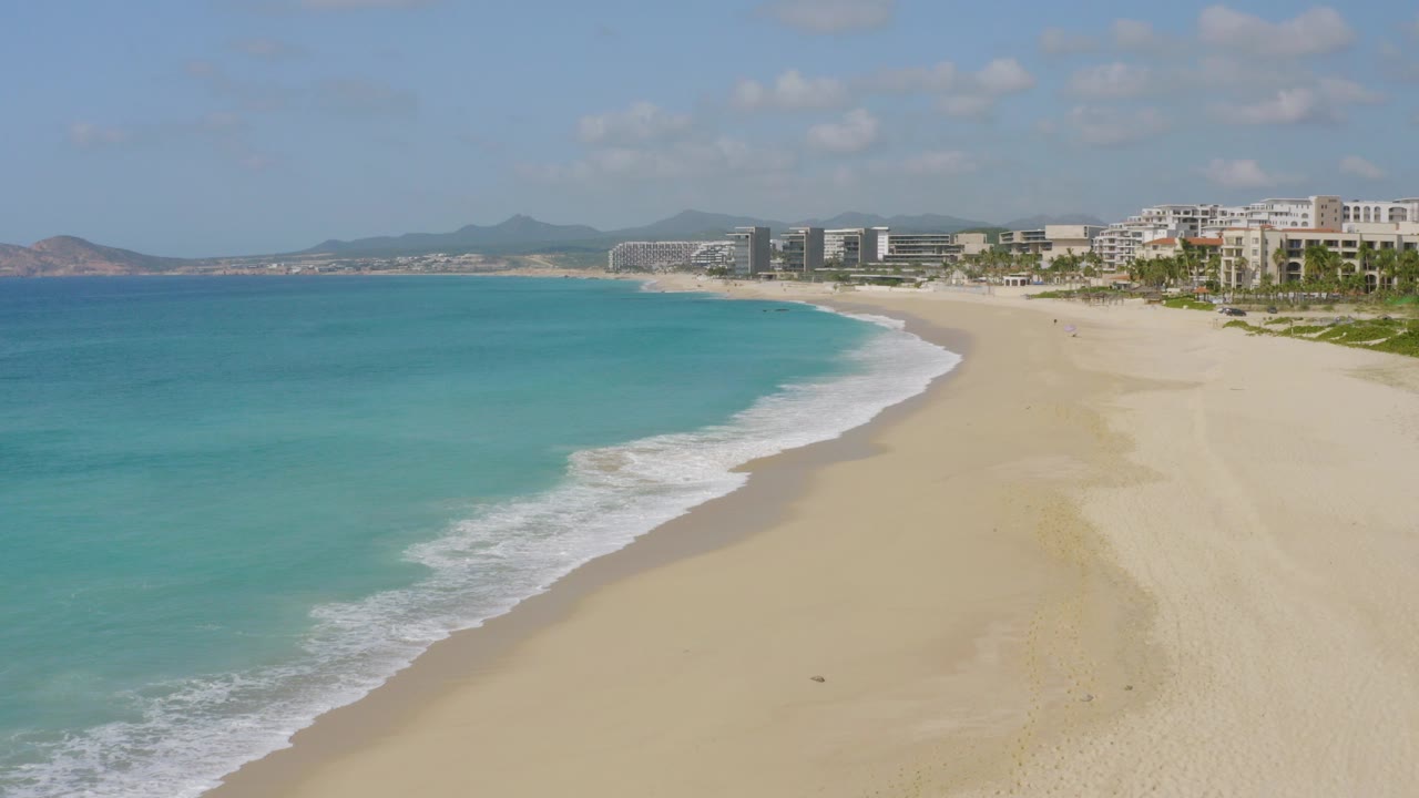 Opening frame of Dreams Beach in Baja California Sur, Mexico, with turquoise water on the left, pale sand on the right, resort buildings in the distance, and no visible people.