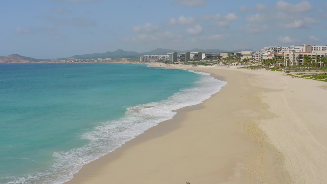 Aerial view over Dreams Beach, Baja California Sur, Mexico, showing blue ocean, a foamy wave edge, pale beach, background buildings, and no visible people.