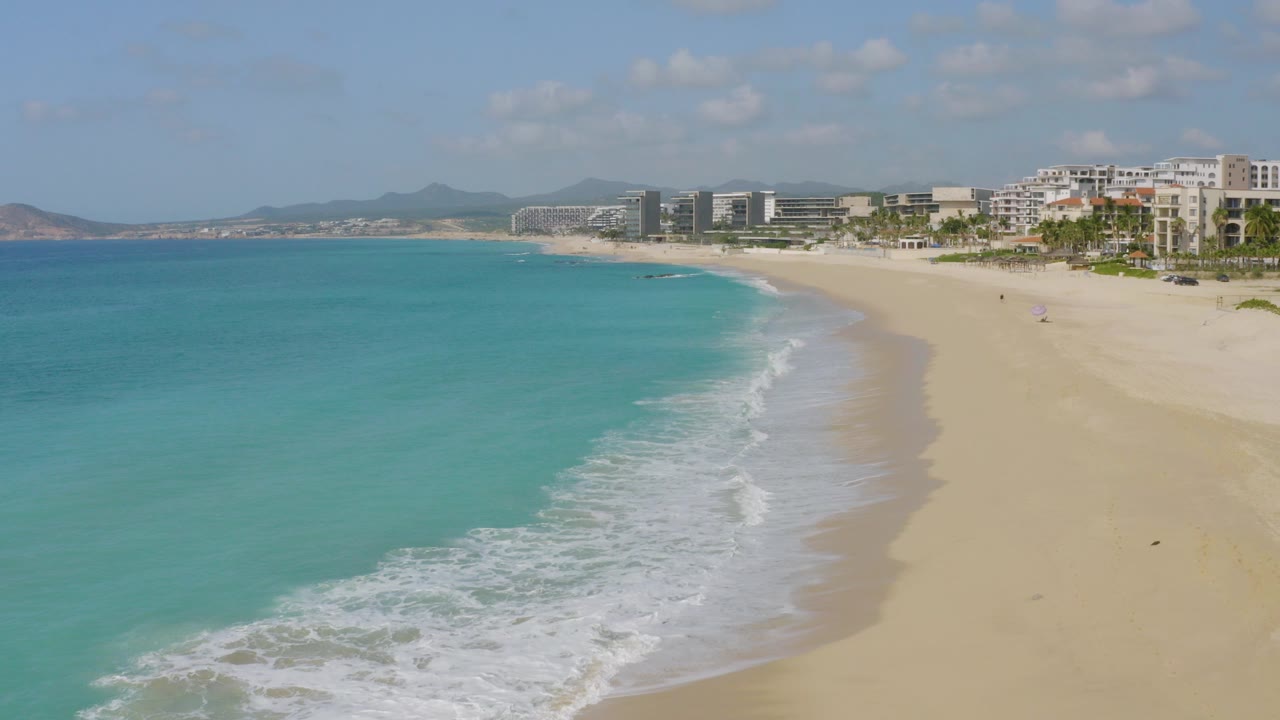 Aerial frame over Dreams Beach, Baja California Sur, with turquoise ocean at left, white surf on pale sand, beachfront buildings and palms at right, and no visible people.