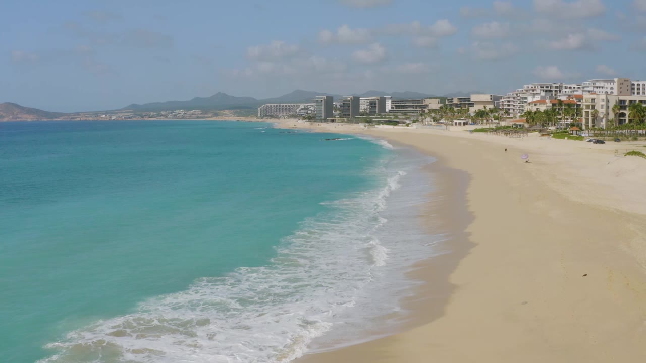 Aerial view at Dreams Beach, Baja California Sur, showing a longer strip of foam along the shore, open sand to the right, modern buildings beyond, and no visible people.