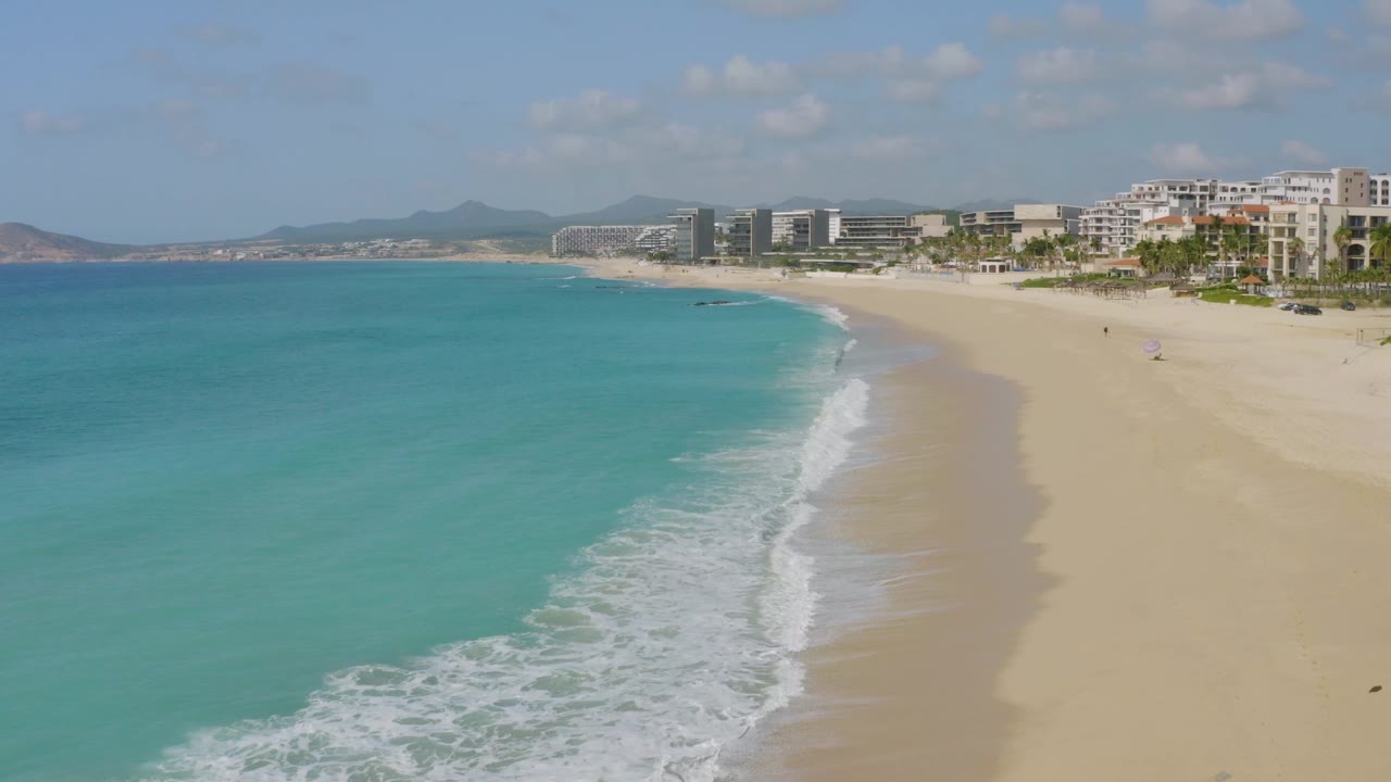 Aerial frame from Dreams Beach, Baja California Sur, with the surf band running diagonally near center, broad sand on the right, turquoise water on the left, and no visible people.