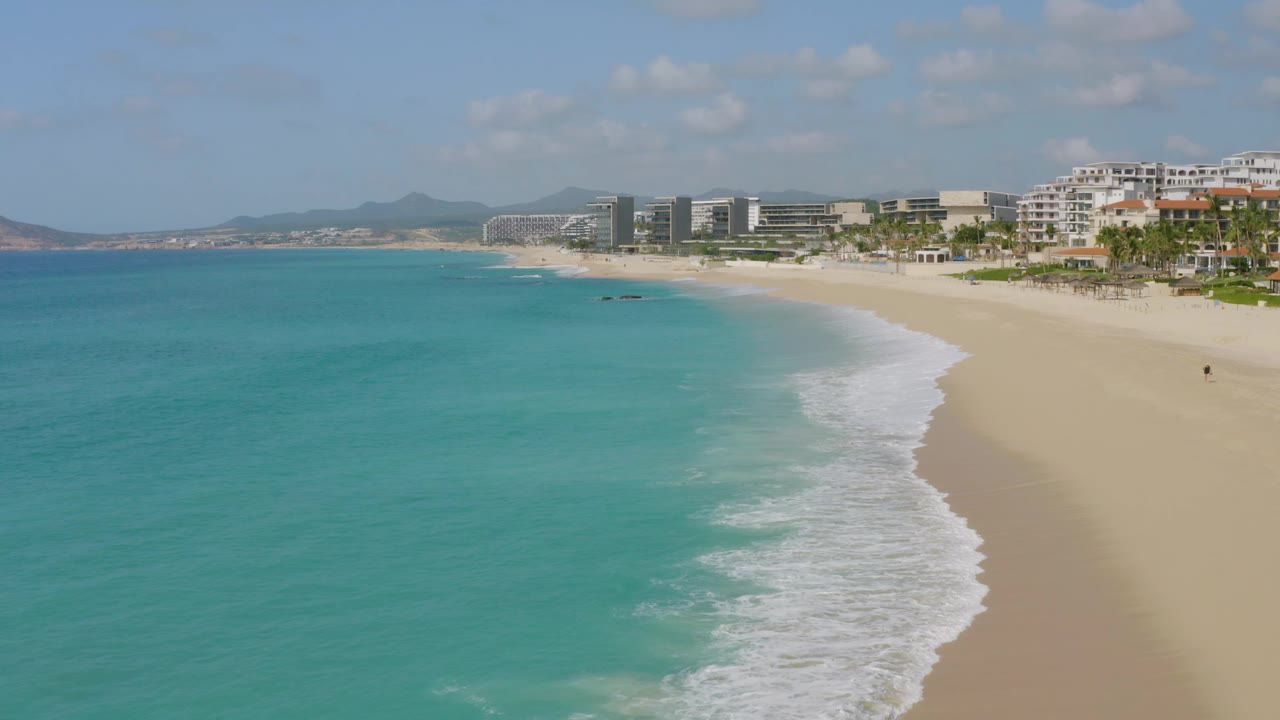 High view over Dreams Beach in Baja California Sur, Mexico, with blue-green water spreading across the left, a curving foamy shoreline, tan sand, resort-style buildings, and no visible people.