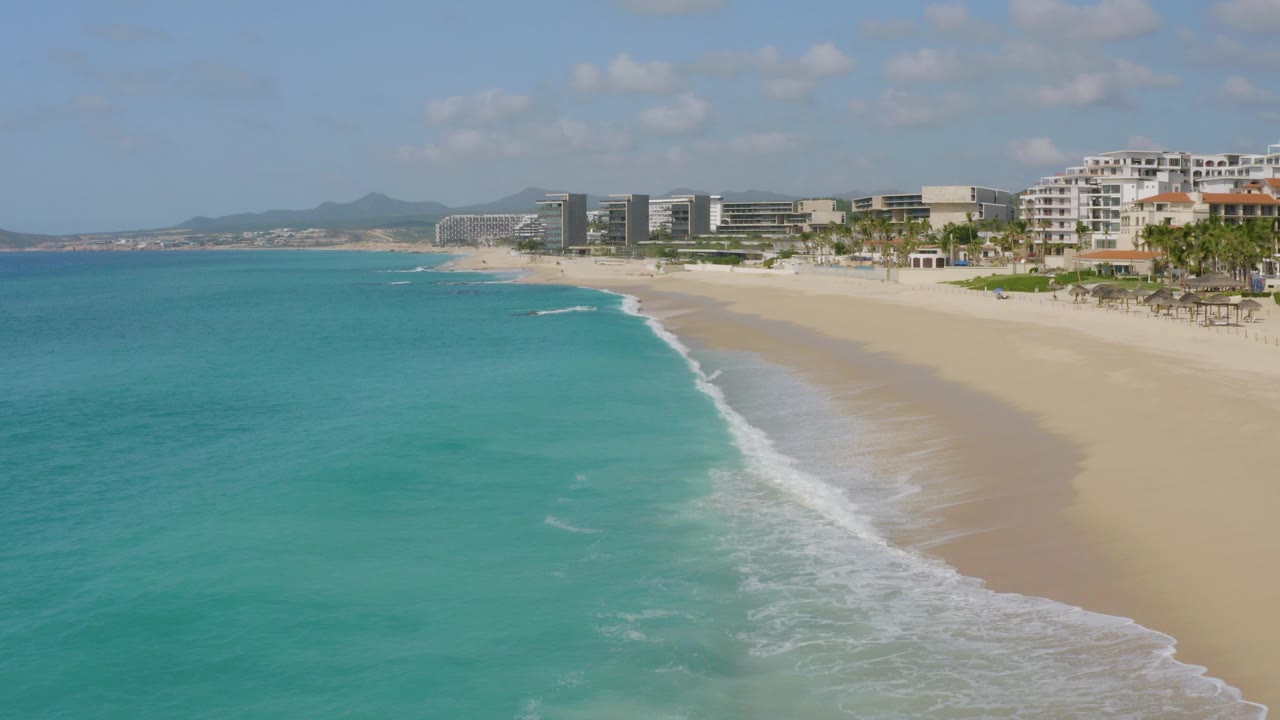 Aerial frame over Dreams Beach in Baja California Sur showing turquoise ocean on the left, pale sand and surf in the center, resort buildings on the right, distant hills, and no visible people.
