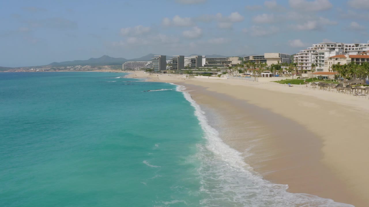 Aerial frame of Dreams Beach showing a stronger diagonal surf line, open turquoise water across the left half, pale sand on the right, resort buildings near the horizon, and no visible people.