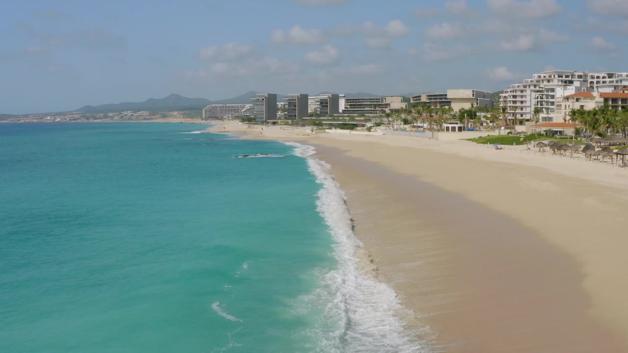 Aerial beach view with turquoise water, white surf, sand, and distant hills