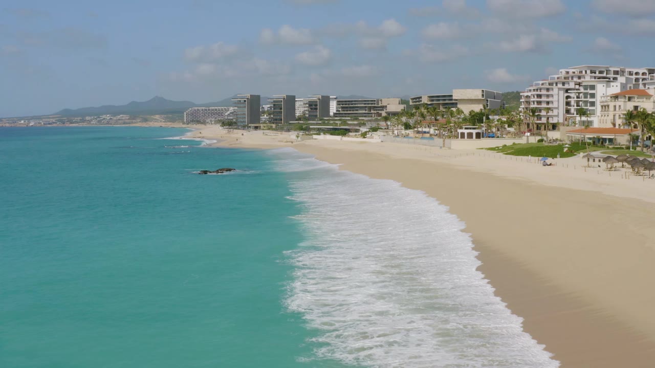 One second into the clip, foamy surf pushes farther up the sand at Dreams Beach while turquoise water fills the left side and beachfront buildings line the upper right, with no visible people.