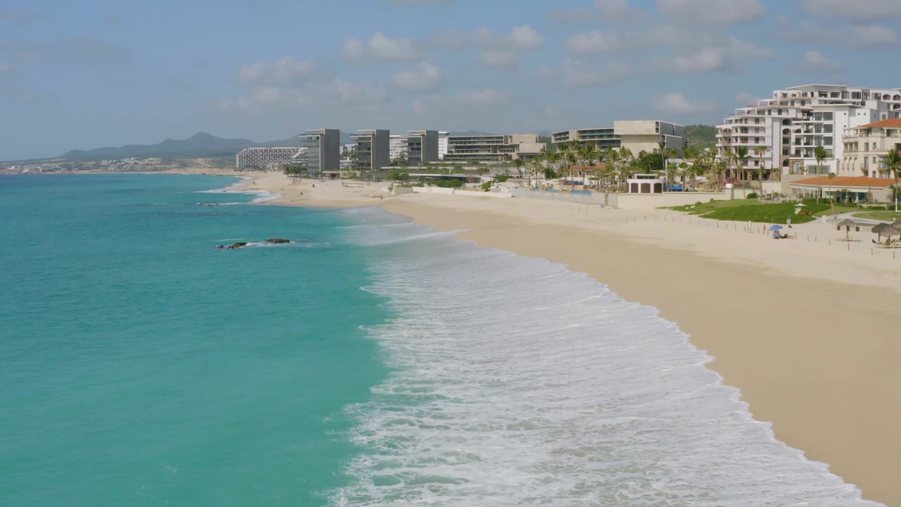 At 0:03, the aerial view shows more open sand on Dreams Beach, a diagonal edge of white surf, turquoise water at left, and modern buildings set back from the shoreline, with no visible people.
