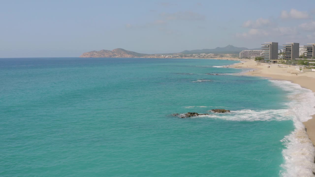 At 0:00, an elevated view of Dreams Beach in Baja California Sur shows turquoise water in the foreground, surf on nearshore rocks, a sandy beach, modern buildings to the right, distant hills, and no visible people.
